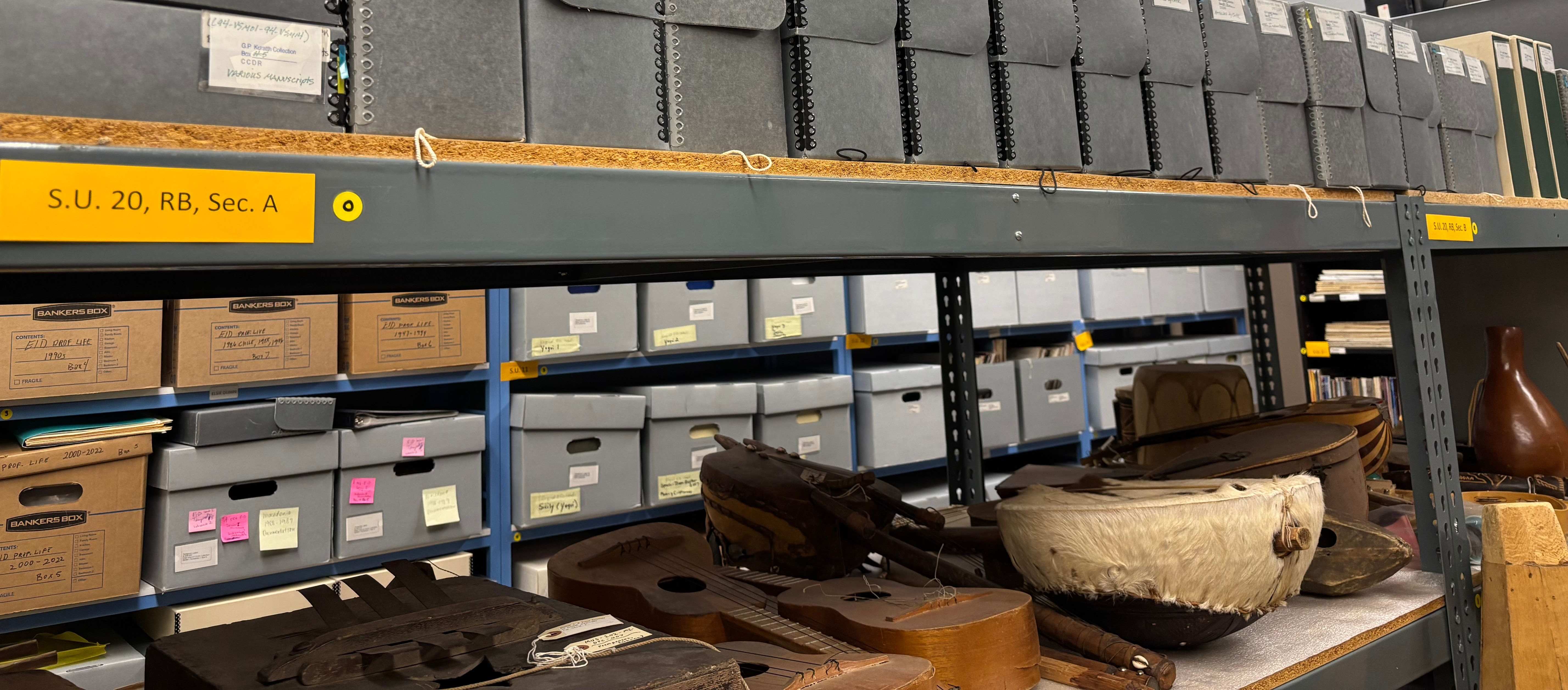 A color photograph of an archival storage room with metal shelving units filled with labeled gray document boxes and binders. The shelves extend across the frame under fluorescent lighting. On a lower shelf in the foreground are musical instruments and objects, including wooden stringed instruments and a drum with a stretched hide surface. The image emphasizes organized archival storage, material culture, and the preservation of performance related collections.