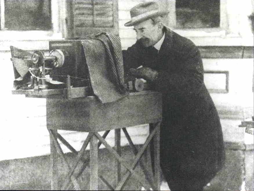 A man wearing a fedora and a long dark coat stands outside next to a white house, working a bellows camera.