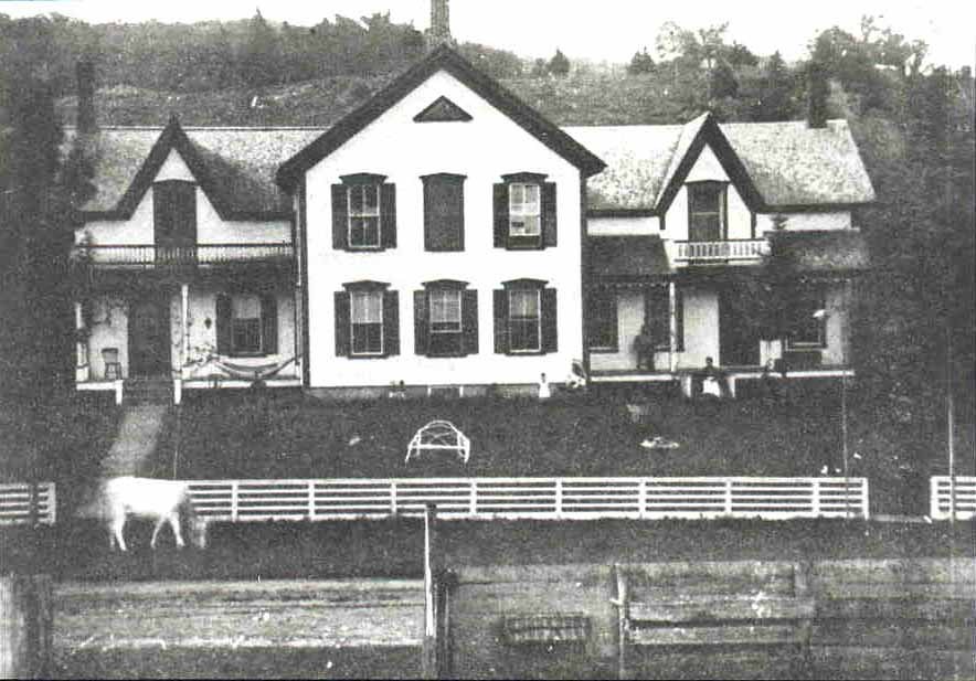 A cow grazes in front of a rural manor with symmetrical second-floor balconies and a long front porch.