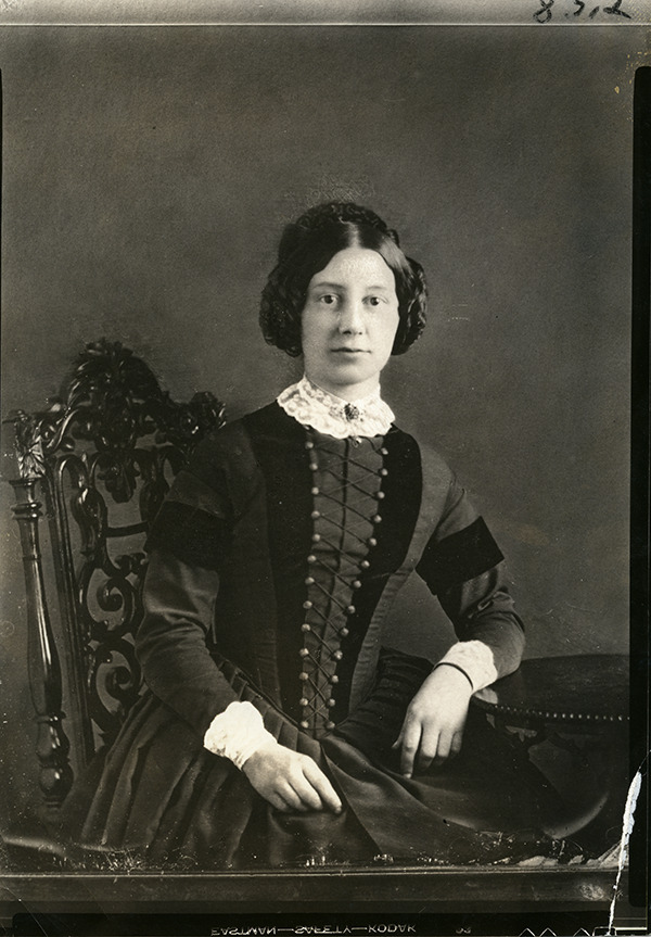 A black and white photograph of Hanna Jane Langstroth Drexel seated in a wooden chair with her left arm propped on a table. She is wearing a dark long-sleeved dress with a white lace collar, and her dark hair is curled and pinned up.