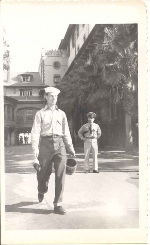 Blurry black and white photo of a walking Coast Guardsman with a paintbrush in one hand and a bucket in the other while an officer watches him.