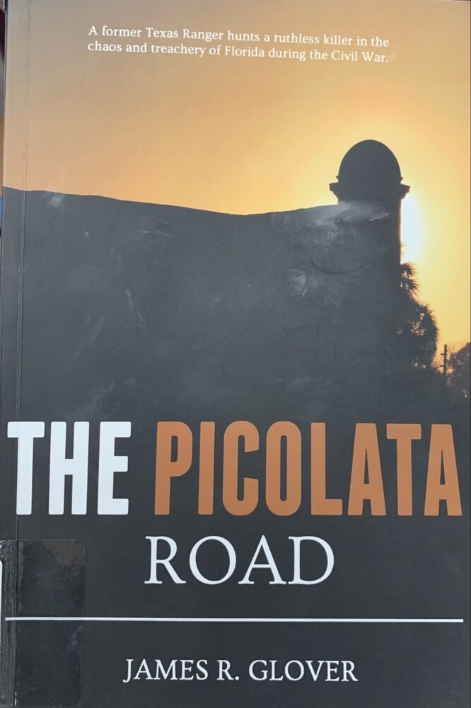 This book is titled "The Picolata Road" by James R. Glover. The book cover is a picture of the Castillo de San Marcos during sunset. The sun is behind one of the towers.