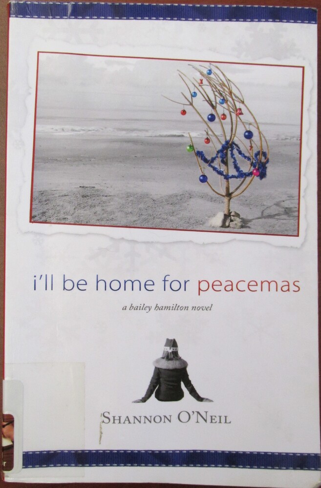 This book is titled "I'll Be Home for Peacemas" by Shannon O'Neil. The book cover is white and includes a photograph of a stick stuck in sand at the beach and decorated like a Christmas tree.