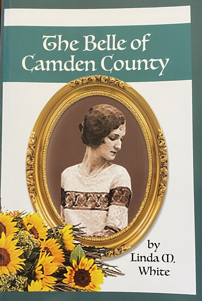 This book it titled "The Belle of Camden County" by Linda M. White. The center of this book cover is an oval portrait of a young woman with a short haircut, looking down and to the right. Her portrait is in a gold, oval frame. On the bottom left corner of the cover, there is a bouquet of sunflowers. The title of the book sits above her portrait in white lettering.