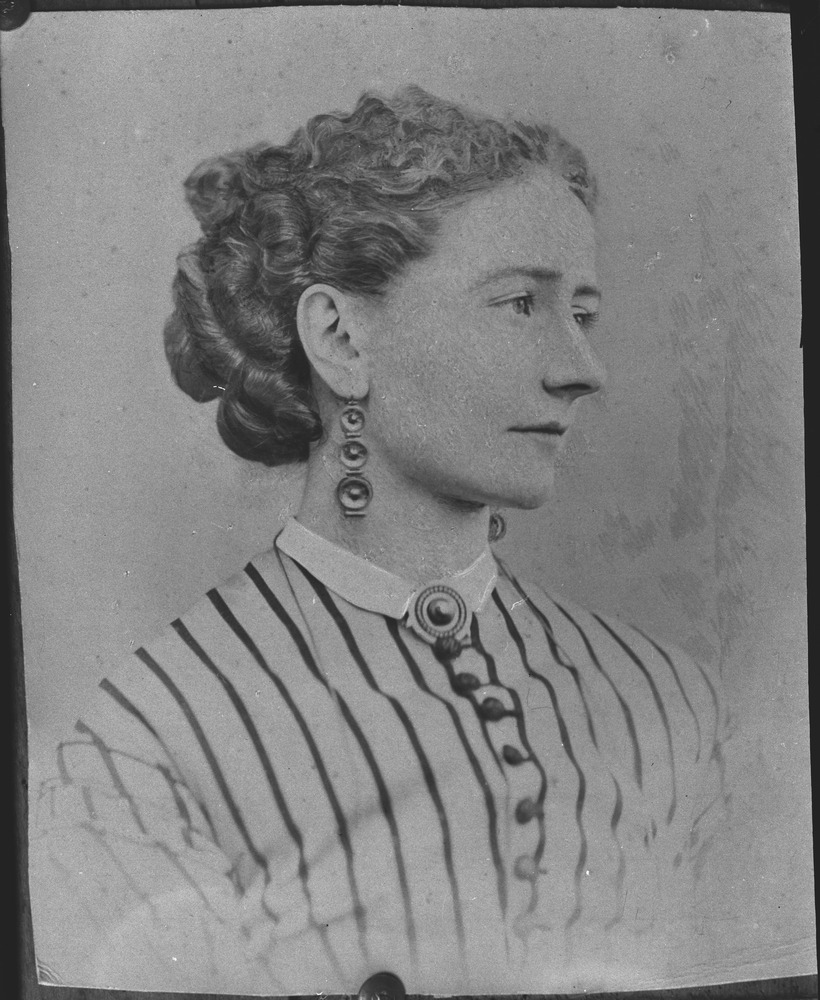 A black and white photograph of a young woman's head and shoulders. Her head is turned in side profile. Her gaze is soft and her expression is neutral. Her curled hair is loosely swept back and neatly coiled and pinned at the back of her head. She is wearing earrings with a drop of three circular shapes getting larger in size with the largest at the bottom. She wears pale clothing with dark narrow vertical lines and a subtle ruffled trim at the shoulders. A row of dark buttons runs down the front and a slim white collar is decorated with a circular brooch similar in design to her earrings.